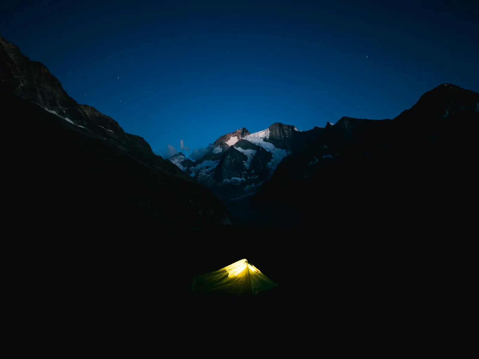 Glowing tent camped beneath dark mountain peaks under a starry night sky