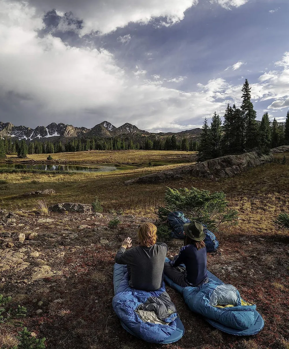 Two campers in sleeping bags looking out over a mountain lake at dawn